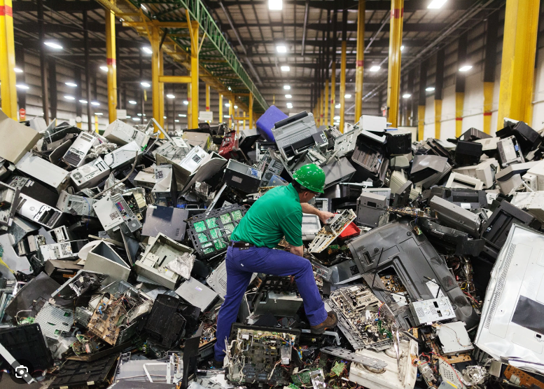 A warehouse worker wearing a green hard hat, green shirt and blue pants stands inside a warehouse on a mountain of broke computers and other electronic equipment inspecting it