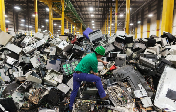 A warehouse worker wearing a green hard hat, green shirt and blue pants stands inside a warehouse on a mountain of broke computers and other electronic equipment inspecting it